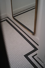 A corner of a room featuring a large mirror with a golden frame mounted on a white wall. The floor is tiled with a white grid pattern, accented by black lines forming geometric shapes.