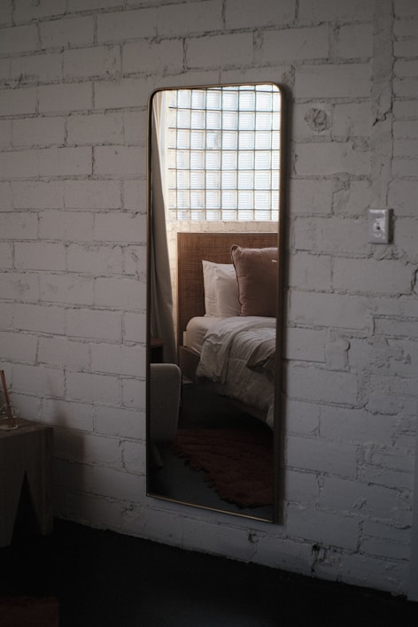 A tall mirror is mounted on a white brick wall, reflecting the image of a bedroom with a bed, pillows, and a window with grid-patterned glass blocks. The setting suggests a modern, cozy interior with a combination of rustic and contemporary design elements.