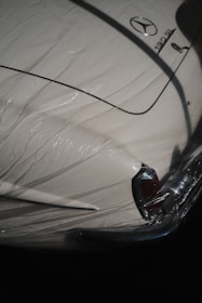 A classic car partially covered by a protective plastic wrap, showcasing a vintage design with a distinctive tail fin. The car is cream-colored with shiny chrome details and a recognizable emblem on the trunk.