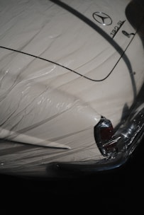 A classic car partially covered by a protective plastic wrap, showcasing a vintage design with a distinctive tail fin. The car is cream-colored with shiny chrome details and a recognizable emblem on the trunk.