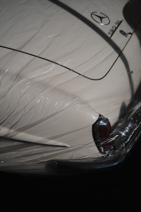 A classic car partially covered by a protective plastic wrap, showcasing a vintage design with a distinctive tail fin. The car is cream-colored with shiny chrome details and a recognizable emblem on the trunk.