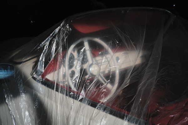 A steering wheel wrapped in a translucent plastic cover, viewed inside a vehicle. The image has a dark background, and the light reflects off the plastic, creating a glossy effect.