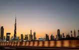 A city skyline with a prominent, tall skyscraper, likely the Burj Khalifa, seen at dusk. The sky transitions from a warm orange near the horizon to a deep blue overhead, creating a serene backdrop for the modern cityscape. The buildings are illuminated, their lights twinkling against the evening sky.