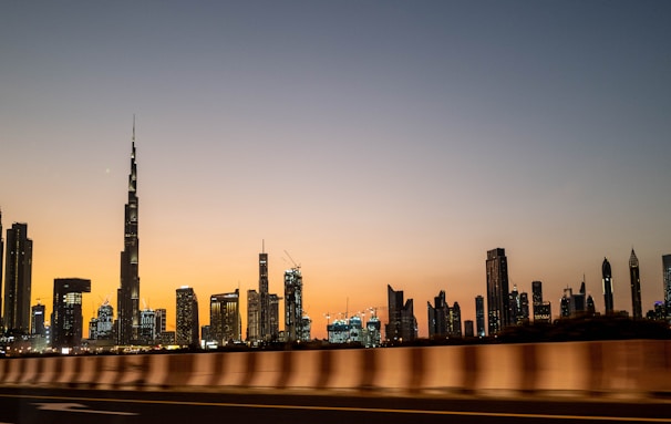 A city skyline with a prominent, tall skyscraper, likely the Burj Khalifa, seen at dusk. The sky transitions from a warm orange near the horizon to a deep blue overhead, creating a serene backdrop for the modern cityscape. The buildings are illuminated, their lights twinkling against the evening sky.