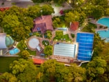 This aerial view captures a large swimming pool complex surrounded by greenery. There are multiple outdoor and indoor pools, including a rectangular swimming pool with lanes, a circular pool, and smaller pools. Buildings with red-tiled roofs are interspersed with the pools, and pathways connect different areas. The entire area is surrounded by dense trees and foliage, providing a tranquil and secluded setting.