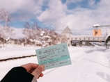 A person is holding a train ticket with snowy scenery in the background. The ground and trees are covered with snow, suggesting a cold winter day. A bridge or overpass is visible in the distance, adding depth to the scene.