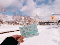A person is holding a train ticket with snowy scenery in the background. The ground and trees are covered with snow, suggesting a cold winter day. A bridge or overpass is visible in the distance, adding depth to the scene.