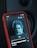 A smartphone with a red case displaying a music player interface. The screen shows an image of a person with blue-tinted hair and beard. Below the image, song information reads 'Is There Someone Else?' by The Weeknd. A speaker or dark surface is visible in the background.