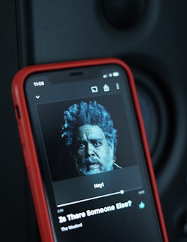 A smartphone with a red case displaying a music player interface. The screen shows an image of a person with blue-tinted hair and beard. Below the image, song information reads 'Is There Someone Else?' by The Weeknd. A speaker or dark surface is visible in the background.