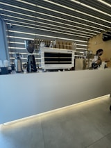 A modern coffee shop counter with a sleek espresso machine, coffee grinder, and stacks of paper cups. The ceiling features a striped pattern with prominent lighting.