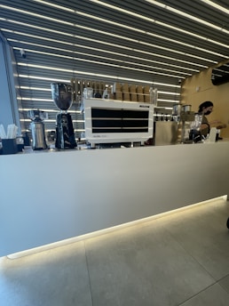 A modern coffee shop counter with a sleek espresso machine, coffee grinder, and stacks of paper cups. The ceiling features a striped pattern with prominent lighting.