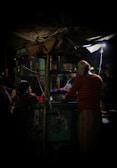 A dimly lit street food stall at night, where a vendor wearing a striped shirt is preparing food. A small group of people, including a child, is gathered around the stall waiting for their order. The scene is illuminated by a single overhead light bulb, creating a cozy and intimate ambiance.