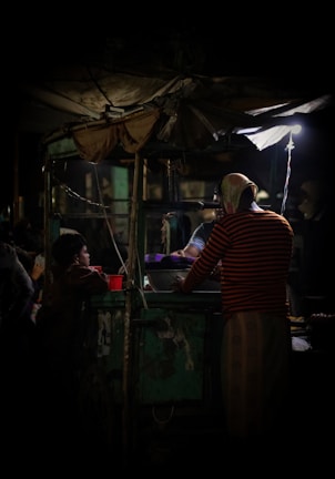 A dimly lit street food stall at night, where a vendor wearing a striped shirt is preparing food. A small group of people, including a child, is gathered around the stall waiting for their order. The scene is illuminated by a single overhead light bulb, creating a cozy and intimate ambiance.
