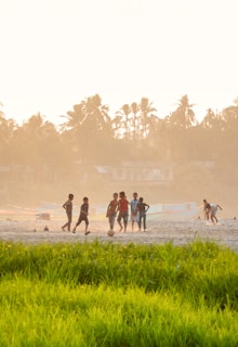 A group of children playing soccer on a sandy field surrounded by lush green grass. In the background, there are silhouettes of palm trees and a few small structures, with a warm, hazy sky overhead.