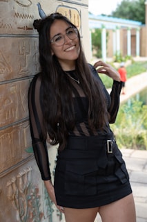 A woman with long dark hair and glasses stands smiling next to a wall adorned with Egyptian-style hieroglyphs. She is wearing a black dress with a transparent striped pattern on the top and a belt around the waist. In the background, there is a garden area with greenery and a person wearing a red shirt.