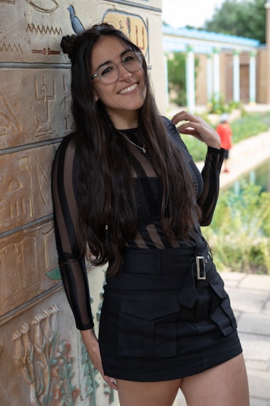 A woman with long dark hair and glasses stands smiling next to a wall adorned with Egyptian-style hieroglyphs. She is wearing a black dress with a transparent striped pattern on the top and a belt around the waist. In the background, there is a garden area with greenery and a person wearing a red shirt.