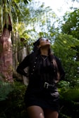 A person with long dark hair stands in a lush green environment, looking upwards. Tropical plants and ferns surround the individual, while sunlight filters through the leaves, creating a vibrant and serene atmosphere.