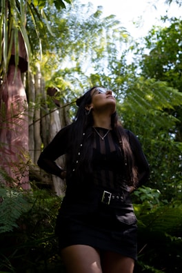 A person with long dark hair stands in a lush green environment, looking upwards. Tropical plants and ferns surround the individual, while sunlight filters through the leaves, creating a vibrant and serene atmosphere.