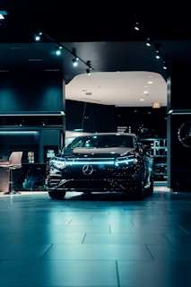 A sleek, modern black car is prominently displayed in a dimly lit showroom with spotlights highlighting its design. The showroom features a polished tiled floor and contemporary furnishings.