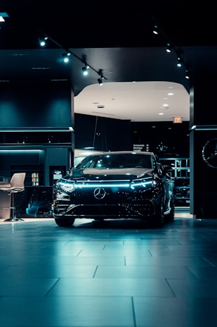 A sleek, modern black car is prominently displayed in a dimly lit showroom with spotlights highlighting its design. The showroom features a polished tiled floor and contemporary furnishings.