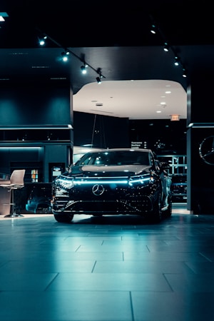 A sleek, modern black car is prominently displayed in a dimly lit showroom with spotlights highlighting its design. The showroom features a polished tiled floor and contemporary furnishings.