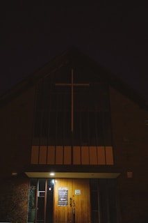 A dimly lit church entrance with a large cross above the main door, illuminated by a small light. The building appears to be made of brick with narrow, tall windows.