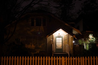A small house with a warmly lit porch surrounded by darkness. The house number 4306 is visible above the door, which has a detailed glass window. In front of the house, there is a white picket fence. Architecture is traditional and simple, creating a cozy atmosphere.