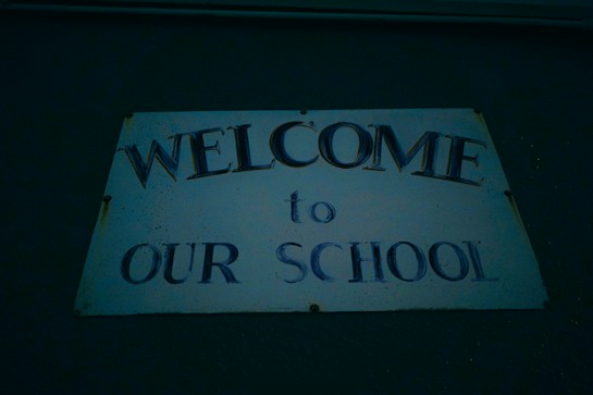 A sign with text that reads 'Welcome to Our School' mounted on a wall. The text is in a bold font, and the sign appears to be weathered with some rust spots at the edges.