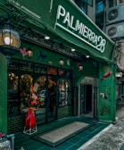 A vibrant storefront with a green facade featuring artistic illustrations of birds and foliage. The entrance is adorned with a flower arrangement in a red wrapper, standing by a set of glass doors. A large illuminated sign displaying 'PALMIERRA28' stands prominently above the entrance. The building has a classic, inviting aesthetic with hanging lanterns contributing to an ambient atmosphere.