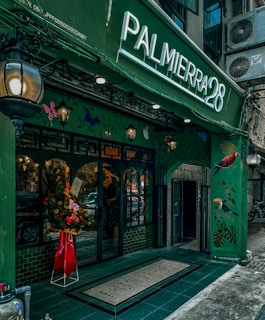 A vibrant storefront with a green facade featuring artistic illustrations of birds and foliage. The entrance is adorned with a flower arrangement in a red wrapper, standing by a set of glass doors. A large illuminated sign displaying 'PALMIERRA28' stands prominently above the entrance. The building has a classic, inviting aesthetic with hanging lanterns contributing to an ambient atmosphere.