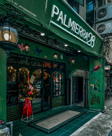 A vibrant storefront with a green facade featuring artistic illustrations of birds and foliage. The entrance is adorned with a flower arrangement in a red wrapper, standing by a set of glass doors. A large illuminated sign displaying 'PALMIERRA28' stands prominently above the entrance. The building has a classic, inviting aesthetic with hanging lanterns contributing to an ambient atmosphere.