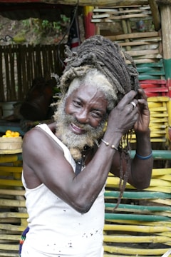 A person with a long beard and dreadlocks is arranging their hair with one hand. They are wearing a white sleeveless top and accessorized with bracelets. The background includes bamboo structures, colorful stripes, and some greenery.