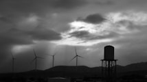 A black and white image depicting a rural landscape with several wind turbines and a prominent water tower. The sky is filled with dramatic clouds, and sunlight breaks through in beams, creating a striking contrast.