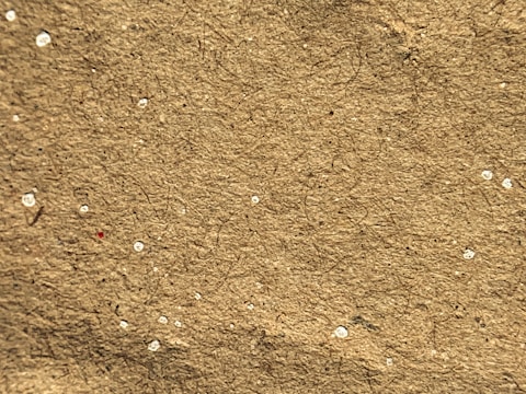 A close-up view of a textured brown surface with small white specks scattered throughout. The material appears fibrous, suggesting it might be a piece of cardboard or recycled paper.