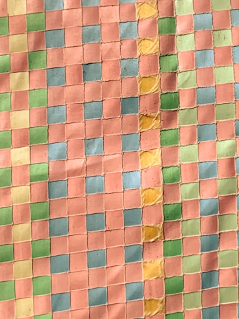 A detailed mosaic pattern made up of small square tiles in a grid layout. The tiles are predominantly pink, with interspersed rows and columns of green, blue, yellow, and light green tiles. There is a visible slight curve in the arrangement of the tiles, creating an artistic effect.