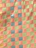 A close-up view of a woven mat made of colorful small squares arranged in a checkerboard pattern. The squares are pastel-colored, including shades of pink, green, and blue, creating a visually engaging texture with slight reflections indicating a shiny surface.