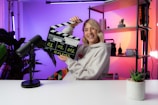 A person smiling and holding a clapperboard, seated at a table with a microphone. The background features colorful gradient lighting and shelving with decorative items and plants.