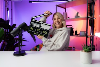 A person smiling and holding a clapperboard, seated at a table with a microphone. The background features colorful gradient lighting and shelving with decorative items and plants.