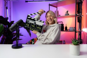 A person smiling and holding a clapperboard, seated at a table with a microphone. The background features colorful gradient lighting and shelving with decorative items and plants.