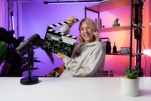 A person smiling and holding a clapperboard, seated at a table with a microphone. The background features colorful gradient lighting and shelving with decorative items and plants.