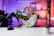A person smiling and holding a clapperboard, seated at a table with a microphone. The background features colorful gradient lighting and shelving with decorative items and plants.