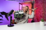A person is sitting at a desk in a brightly lit studio environment, holding a clapperboard with the other hand raised in a gesture. There are colorful lights with gradients of orange, pink, and purple in the background. A microphone is placed on the table next to a small potted plant.