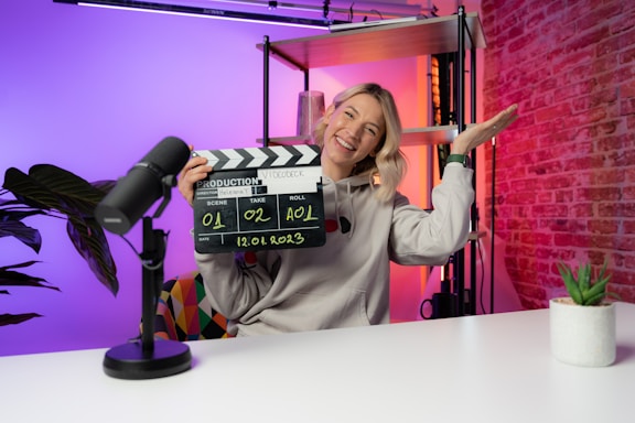 A person is sitting at a desk in a brightly lit studio environment, holding a clapperboard with the other hand raised in a gesture. There are colorful lights with gradients of orange, pink, and purple in the background. A microphone is placed on the table next to a small potted plant.
