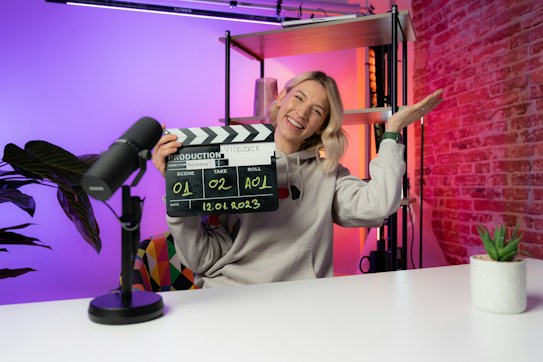A person is sitting at a desk in a brightly lit studio environment, holding a clapperboard with the other hand raised in a gesture. There are colorful lights with gradients of orange, pink, and purple in the background. A microphone is placed on the table next to a small potted plant.
