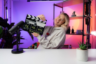 A person wearing a hoodie is smiling while holding a film clapperboard labeled with various details such as scene, take, roll, and date. The scene is set in a brightly lit room with colorful lighting in shades of purple and orange. A microphone is placed on the white desk in front, and there are shelves with decorative items like a plant and figurines in the background.