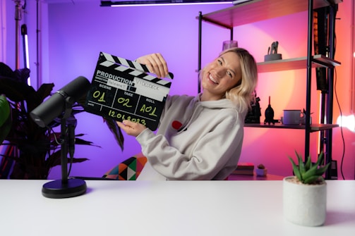 A person wearing a hoodie is smiling while holding a film clapperboard labeled with various details such as scene, take, roll, and date. The scene is set in a brightly lit room with colorful lighting in shades of purple and orange. A microphone is placed on the white desk in front, and there are shelves with decorative items like a plant and figurines in the background.