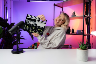 A person wearing a hoodie is smiling while holding a film clapperboard labeled with various details such as scene, take, roll, and date. The scene is set in a brightly lit room with colorful lighting in shades of purple and orange. A microphone is placed on the white desk in front, and there are shelves with decorative items like a plant and figurines in the background.