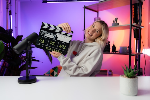 A person wearing a hoodie is smiling while holding a film clapperboard labeled with various details such as scene, take, roll, and date. The scene is set in a brightly lit room with colorful lighting in shades of purple and orange. A microphone is placed on the white desk in front, and there are shelves with decorative items like a plant and figurines in the background.