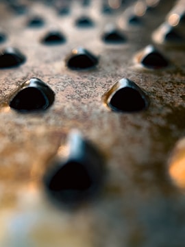 A close-up view of a metallic surface with numerous small, pyramid-shaped protrusions. The texture appears rough, with a series of repeating patterns that create an abstract, industrial feel.