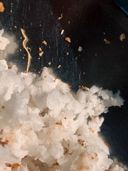 A close-up view of shredded, white coconut, with small brown specks scattered throughout. The background is dark, emphasizing the texture and color variation within the coconut pieces.